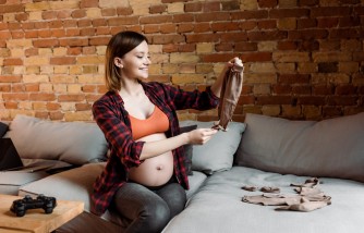 cheerful and pregnant woman holding baby pants near joystick on table 