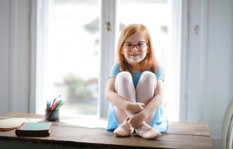 Happy cute small girl in glasses sitting on table in light living room