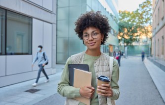 Beautiful curly female student holds notebook digital tablet and takeaway coffee focused into distance poses in urban setting wears casual clothes poses at busy street being on way to university
