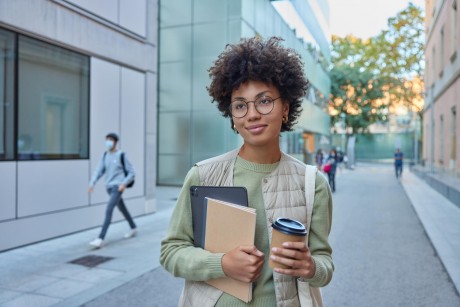 Beautiful curly female student holds notebook digital tablet and takeaway coffee focused into distance poses in urban setting wears casual clothes poses at busy street being on way to university